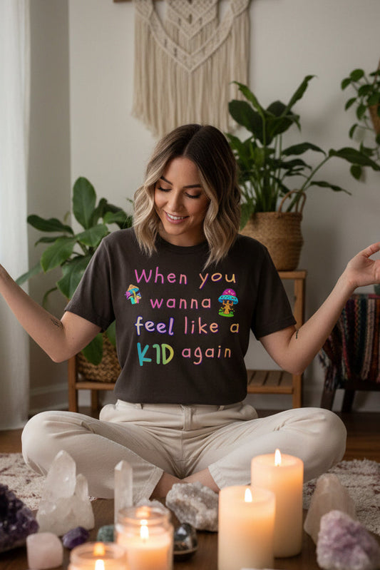 A dark brown short-sleeve t-shirt with a colorful graphic text print that reads 'When you wanna feel like a KID again' in pastel rainbow colors, accompanied by two small cartoon figures, shown on a plain white background.