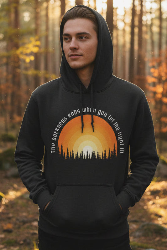 A dark charcoal gray pullover hoodie with a front kangaroo pocket and a graphic print of a sunset over a pine tree silhouette. The text 'The darkness ends when you let the light in' is written in white around the sun graphic.