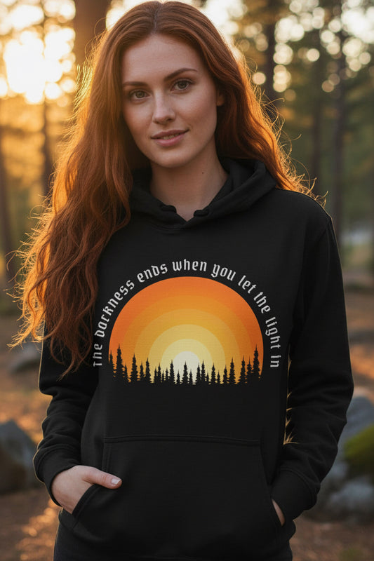 A black pullover hoodie with a sunset graphic on the chest. The graphic features a yellow, orange, and red gradient sun with black pine tree silhouettes and white curved text that reads, 'The darkness ends when you let the light in.
